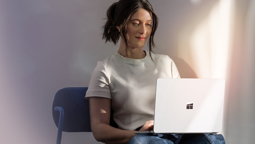 A woman sitting in a blue chair using a Surface Laptop 7.