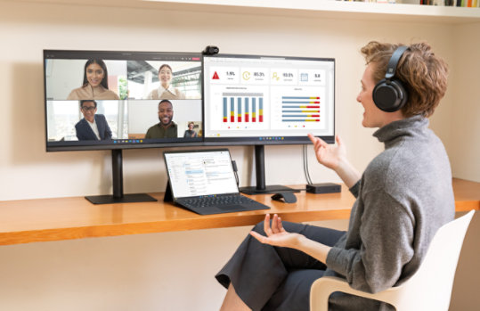  A woman sitting at a home office desk working on a Surface Pro 9 showing Outlook on a Teams call and connected to two external monitors showing Dynamics 365.